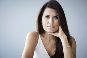San Diego patient with long dark hair, wearing a white top, rests her chin on her hand and looks at the camera during facelift recovery.